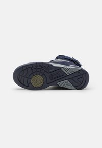 Semelle d'une basket bleu marine avec des accents gris, un logo circulaire portant l'inscription « Patrick Ewing » et le nom de la marque « Ewing » embossé sur la semelle.