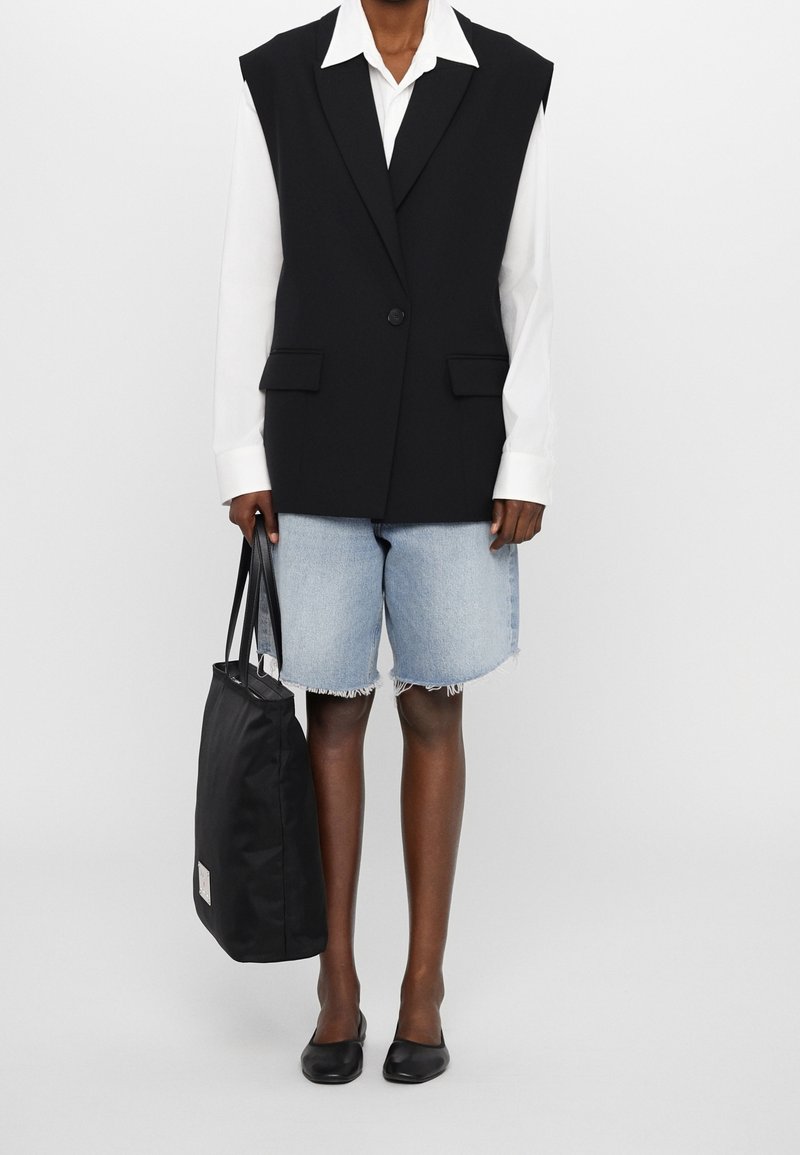 Personne portant un blazer noir sans manches sur une chemise blanche à manches longues, un short en jean bleu clair, des ballerines noires, et portant un sac cabas noir.