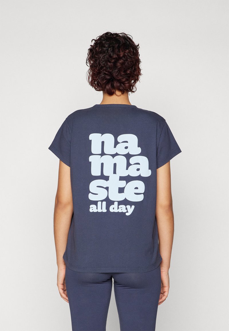 Tamno plava pamučna majica s kratkim rukavima. Stražnja strana sadrži veliki, svijetloplavi natpis "namaste all day" u podebljanom, zaobljenom fontu.
