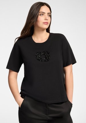 Frau mit langen dunklen Haaren, die ein schwarzes T-Shirt mit einem kleinen schwarzen Perlenblumen-Design trägt, Hände in den Taschen der schwarzen Hose, schaut nach rechts.
