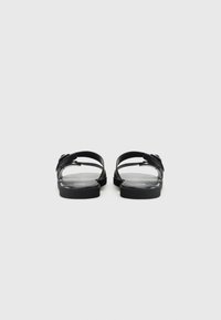 Sandales noires avec une semelle plate, bout ouvert, deux lanières et quincaillerie métallique. Texture lisse mate avec un design minimaliste.