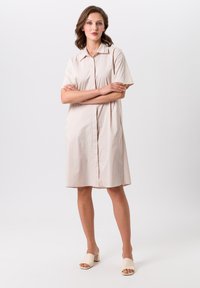 Robe chemise en beige à manches courtes, avec un col classique, une fermeture à boutons sur le devant et une coupe décontractée. Accessoirisée avec des mules à talons carrés de couleur claire.