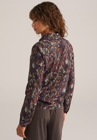 Långärmad blus i lila tyg med abstrakta flerfärgade mönster. Kragsnitt med löst fall och slät textur.