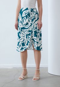 Teal omslagskjørt med hvitt bladmønster, laget av et glatt stoff. Har en sideknute og en frontsplitt for økt bevegelsesfrihet.