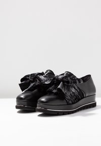 Scarpe nere con una punta in pelle lucida, parte superiore in tessuto nero testurizzato con un grande fiocco decorativo e una spessa suola in gomma a zeppa.