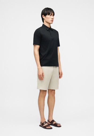 Άνδρας που στέκεται, φοράει μαύρο πόλο μπλουζάκι με κοντά μανίκια, μπεζ βερμούδα μέχρι το γόνατο και σκούρες καφέ δερμάτινες σανδάλες, σε απλή φόντο.