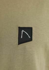 Tessuto verde oliva con una texture liscia, caratterizzato da una toppa rettangolare nera. La toppa presenta un design minimalista bianco.