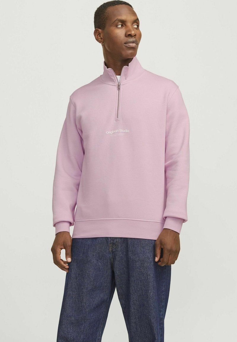 Light pink zip-up sweatshirt with ribbed cuffs and hem, featuring white text "Originals Studio" on the front. Worn with dark denim pants.