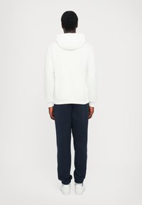 Sweatshirt branco com capuz, com punhos e barra canelados, combinado com calças de jogging azul-marinho e ténis brancos, visto de costas contra um fundo simples.