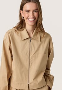 Beige zip-jakke lavet af let materiale, der har en spids krave, en frontlomme og en løs, afslappet pasform med lange ærmer.