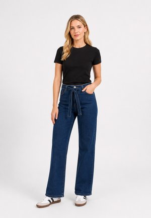 Frau mit langen blonden Haaren, die ein schwarzes kurzärmeliges Shirt, hoch taillierte blaue Jeans mit Bindegürtel und weiße Sneakers mit schwarzen Streifen trägt.