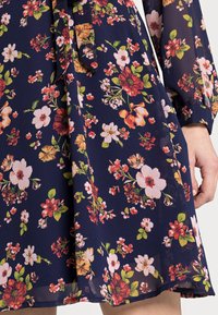 Floral dress in navy blue fabric featuring varied flowers in pink, red, and green. Includes a tie at the neckline and long, defined sleeves.