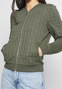 Olivgrön bomberjacka med texturerat mönster, dragkedjestängning, ribbade ärmslut och nederkant, samt sidofickor. Matchas med ljusblå jeans.