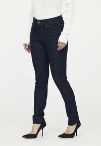 Dünne dunkelblaue Jeans mit orangefarbenen Nähten, die einen hohen Bund und einen Knopfverschluss aufweisen. Kombiniert mit schwarzen High Heels.