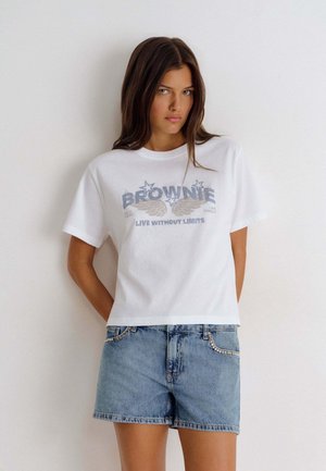 Joven mujer con cabello castaño largo que lleva una camiseta blanca con diseño gráfico y pantalones cortos vaqueros con pedrería, de pie frente a un fondo blanco liso.