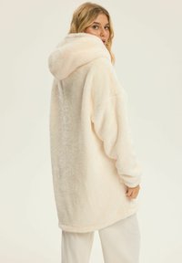 Light beige oversized hoodie made from soft, fluffy material. Features a large hood and loose-fitting sleeves, with rounded hem and texture detailing.