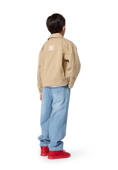 Veste bomber beige avec un patch carré dans le dos, associée à un jean large bleu clair et des baskets rouges. La tenue met en avant une coupe décontractée.