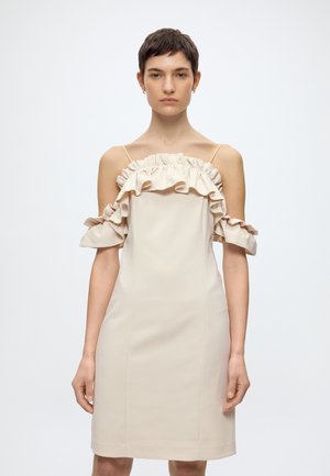 Γυναίκα με αμάνικο μπεζ φόρεμα με βολάν στους ώμους, στέκεται σε λευκό φόντο.