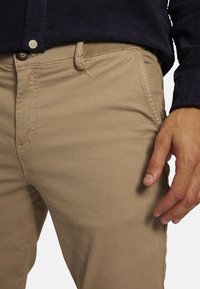 Beige Chinos aus Baumwollmischung mit geradem Schnitt, ausgestattet mit Gürtelschlaufen und Knopfverschluss, neben einem marineblauen Hemd präsentiert.
