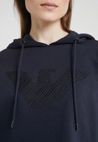Marinblå hoodie med en texturerad, mörk randlogotyp. Har en dragsko på huvan med metallspetsar. Tyget har en jämn, mjuk textur.