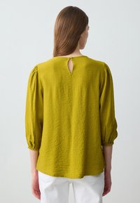 Blusa verde, strutturata, con maniche a palloncino e un'apertura a chiave sul retro, abbinata a pantaloni bianchi.