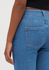 Calças de ganga azuis de cintura alta com um corte slim. Inclui duas bolsos traseiros e uma textura subtil, enfatizando o material clássico da ganga.