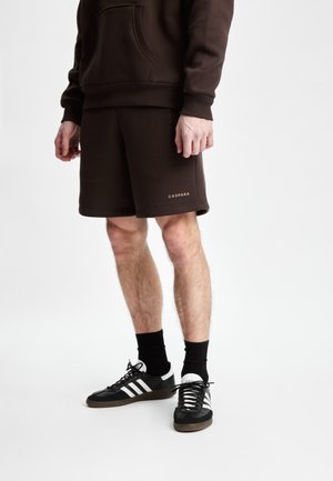 Brun bomull hettegenser med frontlomme, matchende brune shorts, og svarte sneakers med hvite striper, accessorert med svarte sokker.