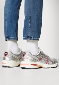 Persona que lleva zapatillas plateadas y rojas con calcetines blancos y vaqueros azules, de pie sobre un suelo blanco contra un fondo liso.