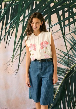 Femme souriante portant un haut blanc sans manches avec broderie florale rose et un pantalon bleu à fines rayures longueur genou, se tenant devant de grandes feuilles de palmier vertes.