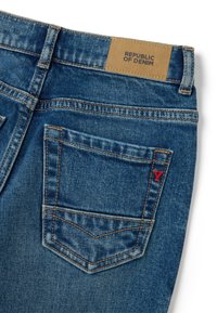 Jeans de mezclilla en un lavado azul medio con costuras naranjas en contraste, con un parche de cuero etiquetado "REPÚBLICA DEL MEZCLILLA" y un detalle de logo rojo.