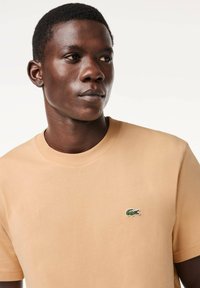 T-shirt in cotone beige chiaro con scollo rotondo, maniche corte e un piccolo logo di coccodrillo verde sul lato sinistro del petto.