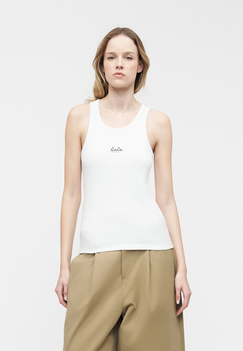 Νεαρή γυναίκα που φοράει λευκό αμάνικο τοπ με μικρό λογότυπο και άνετο μπεζ παντελόνι, στέκεται μπροστά σε ένα απλό φωτεινό φόντο.