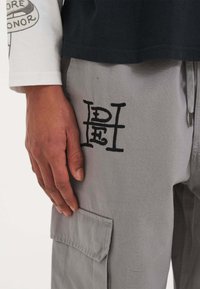 Graue Cargo-Hosen mit einer strukturierten Webart, die ein auffälliges schwarzes gesticktes Logo und eine Seitentasche aufweisen. Eine Hand ruht auf dem Stoff.