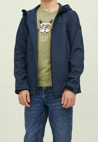 Chaqueta softshell azul marino con capucha, cremallera frontal y puños ajustables, sobre una camiseta gráfica verde claro y vaqueros azules.