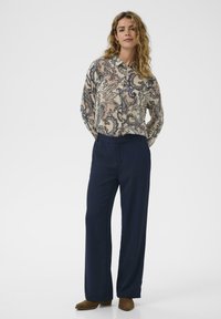Žena stojí v blúzke s paisley vzorom s dlhým rukávom, tmavomodrých nohaviciach s širokými nohavicami a hnedých členkových čižmách na bielom pozadí.