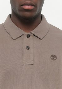 Brown polo shirt made of textured fabric, featuring a two-button placket and a small embroidered logo on the left chest.