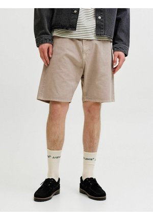 Personne portant un short beige jusqu'aux genoux, des chaussettes blanches de marque, des chaussures noires, une chemise rayée et une veste en jean noire sur fond uni.