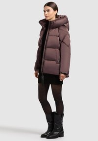 Kvinde iført en mørk mauve pufferjakke med hætte, sort strikkjole, gennemsigtige sorte nylonstrømper og sorte støvler til midt på læggen, som står foran en lys baggrund.