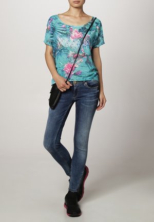 Nainen, jolla on sininen kukkakuvioinen t-paita, tummansiniset skinny-farkut, musta olkalaukku ja mustat lenkkarit, joissa on vaaleanpunaiset pohjat, seisoo jalat ristissä.