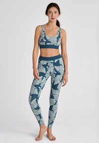 Femme portant un soutien-gorge de sport à motifs floraux teal et blancs et unlegging, debout pieds nus contre un fond blanc.