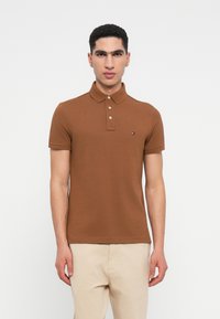 Vīrietis, tērpies brūnā īsām piedurknēm polo kreklā ar trim pogām un bēšos biksēs, stāv uz baltas fona.