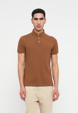 Férfi barna rövidujjú pólót visel, három gombbal, beigem színű nadrággal, háttérben fehér fal.