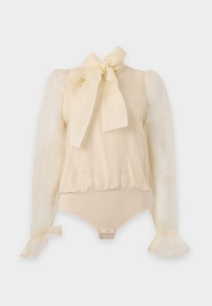 Femme aux longs cheveux foncés portant un chemisier beige transparent avec un nœud, un pantalon gris à jambes larges, une ceinture noire et des sandales noires à talons, debout devant un fond blanc.
