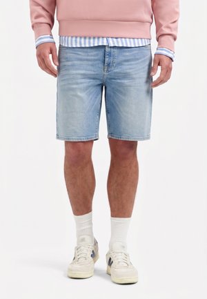 Mann, der hellblaue Jeansshorts, weiße Sneakers, weiße Socken, ein pinkes Sweatshirt und darunter ein blau-weiß gestreiftes Hemd trägt.