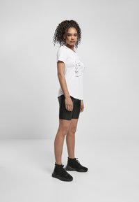 Hvit t-skjorte med en svart linjetegning av et ansikt, kombinert med svarte shorts og lagdelte, teksturerte svarte ankelstøvler.