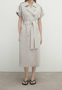 Jasno beżowa sukienka z lnu z krótkimi, podwiniętymi rękawami, podwójnymi guzikami, dopasowanym paskiem w talii oraz prostym, średniej długości krojem.