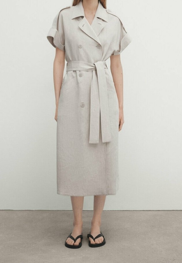 Jasno beżowa sukienka z lnu z krótkimi, podwiniętymi rękawami, podwójnymi guzikami, dopasowanym paskiem w talii oraz prostym, średniej długości krojem.
