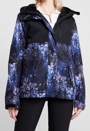 Vrouw draagt een blauw-zwart patroon winterjack met capuchon, borstzak met rits en zijzakken met klep.