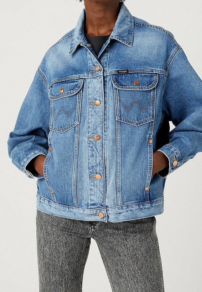 Veste en denim bleu à coupe décontractée, avec deux poches poitrine, des accents en boutons en cuivre et des détails de couture visibles.