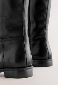 Zbliżenie na czarne skórzane buty z gładką teksturą, detalami przeszyć i solidnymi niskimi obcasami na jasnym tle.